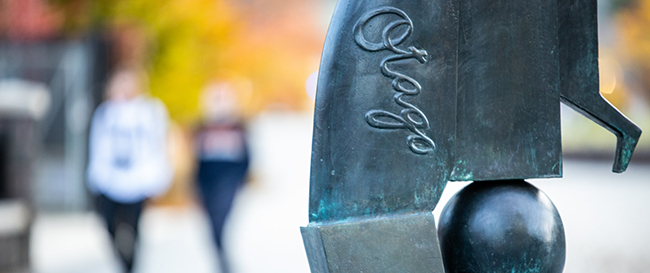 Detail of abstract metal sculpture bearing the word Otago, with people walking out of focus in the background.